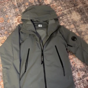 C.P. Company jacka  - Snygg grön vindjacka från C.P. Company med huva och svarta dragkedjor. Jackan har två snedställda fickor framtill och en ikonisk linsdetalj på ärmen. Perfekt för dig som gillar stilren och funktionell design.storlek m men passar s 