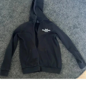 Svart hoodie från Peak Performance - Säljer en svart hoodie från Peak Performance med dragkedja framtill och huva. Tröjan har två fickor och en liten vit logga på bröstet. Perfekt för en avslappnad stil och enkel att matcha med det mesta.