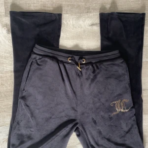 Svarta mjukisbyxor från Juicy Couture - Mysiga svarta mjukisbyxor från Juicy Couture med guldfärgade nitar i loggan på ena benet. Byxorna har resår och snörning i midjan samt fickor både fram och bak. Perfekta för en avslappnad stil.