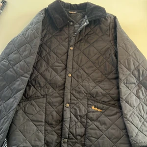 Svart quiltad jacka från Barbour - Klassisk svart quiltad jacka från Barbour med krage i manchester och tryckknappar framtill. Jackan har två stora fickor och broderad Barbour-logga på bröstet. Den är i storleken L (barnstorlek), vilket motsvarar ungefär xxs eller xs (herrstorlek). Ny pris ca 1700 kr och mitt pris 670 kr. Perfekt för kyliga dagar och stilren look. Skriv bara om du har frågor, pris kan diskuteras!