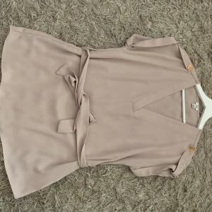 Beige blus med bälte från River Island - Säljer en beige blus från River Island med v-ringning och korta ärmar. Blusen har dekorativa knappar på axlarna och ett matchande bälte i midjan som kan knytas. Perfekt för en stilren och enkel look.