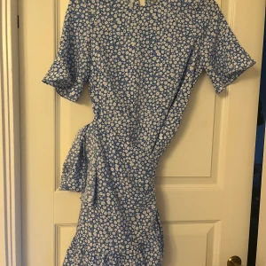 Blå blommig omlottklänning - Söt blå klänning med vitt blommönster och omlottdetalj i midjan. Klänningen har korta ärmar, perfekt för vår och sommar!