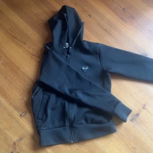 Svart hoodie från Comme des Garçons Play - Svart hoodie med dragkedja från Comme des Garçons Play. Klassisk hjärtlogga med ögon på bröstet och huva med dragsko. Två fickor framtill och ribbade muddar vid ärmslut och nederkant. S men passar Xs, lite liten i storlek