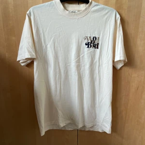 Off white  t-shirt med tryck från Woodbird - Säljer en off white t-shirt med liten svart och beige logga på bröstet från Woodbird.  T-shirten har rund halsringning och är tillverkad i mjuk bomull. Perfekt för en avslappnad stil.