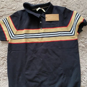 Svart och beige t-shirt från Burberry - Snygg svart t-shirt från Burberry med klassiska beige, vita och röda ränder över bröstet. T-shirten har krage och knappar framtill för en stilren look. Perfekt för dig som vill ha något trendigt och bekvämt.