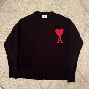 Svart stickad tröja från Ami Paris med rött hjärta - Säljer en svart stickad tröja från Ami Paris med ett stort rött hjärta och bokstaven A broderat på bröstet. Tröjan har rund halsringning och långa ärmar. Perfekt för dig som gillar stilrena plagg med en twist. Helt ny. Kontakta cör mer info o bilder