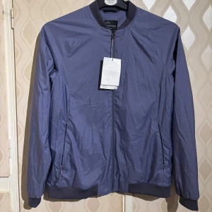 Blå/lila bomberjacka från Peak Performance - Snygg blå bomberjacka från Peak Performance med dragkedja framtill och ribbade muddar vid ärmslut och krage. Jackan har en stilren design och två fickor med dragkedja på sidorna. Perfekt för vår och höst.