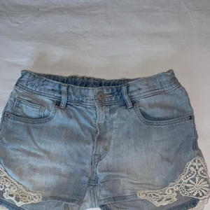Ljusblå jeansshorts med spetsdetaljer - Säljer ett par ljusblå jeansshorts med snygga spetsdetaljer i vitt på sidorna. Klassisk femficksmodell med bälteshällor och knappstängning. Perfekta för varma dagar och en avslappnad stil.