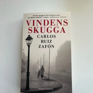 Vindens skugga - En gripande roman av Carlos Ruiz Zafón, utgiven av Norstedts. Boken har ett svartvitt omslag med röd titeltext och ett foto av en gammal stadsgata. Perfekt för dig som gillar mysterier, äventyr och historiska berättelser.