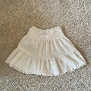 Vit volangkjol från Gina Tricot - Supersöt vit kjol från Gina Tricot med bred smock i midjan och volang nertill. Kjolen är luftig och har en lätt struktur i tyget, perfekt för varma dagar. Köpt 2023 men i bra skick 👍 hör av dig om du har frågor!