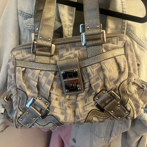 Guess, Beige och grå axelväska - Väska från Guess i beige och grått. Väskan har stora spännen, dragkedja upptill och metallogo framtill. Perfekt som en statementväska💕