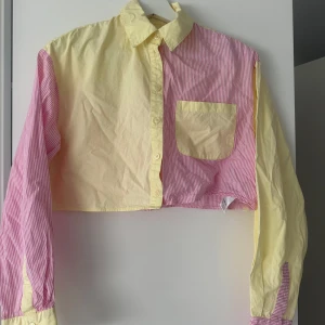 Croppad skjorta i gult och rosa med ränder - Säljer en croppad skjorta med lång ärm i tvådelad design. Ena sidan är ljusgul och andra sidan rosa med vita ränder. Skjortan har en bröstficka och knäppning framtill. Perfekt för dig som gillar färg och unika plagg!