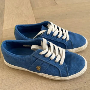 Blå sneakers från Lauren Ralph Lauren - Snygga blå sneakers från Lauren Ralph Lauren med vita snören och vit sula. Skorna har en gul broderad logga på sidan och är tillverkade i textilmaterial. Perfekta för en avslappnad och sportig stil.