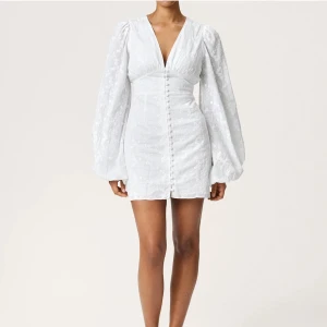 Vit kortklänning med puffärm från Adoore - Supersöt vit kortklänning från Adoore med v-ringning, långa puffärmar och knappar framtill. Aldrig använd med lapparna kvar och kommer med en extra knapp. Nypris 1495kr