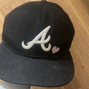New Era keps - Svart 59FIFTY keps från New Era med Atlanta Braves-logga framtill, broderad text och detaljer i vitt och rött. På sidan finns ett gult World Series 1995-märke och texten 'Love & Hustle = Game'. Storlek 7 (55,8 cm).