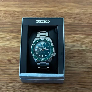Seiko 5 automatisk herrklocka med stållänk - Säljer en Seiko 5 automatisk klocka med grön vridring och svart urtavla. Klockan har dag- och datumvisning samt robust länk i rostfritt stål. Levereras i originalask med certifikat och manual! Klockan har en väldigt liten repa på vridringen!