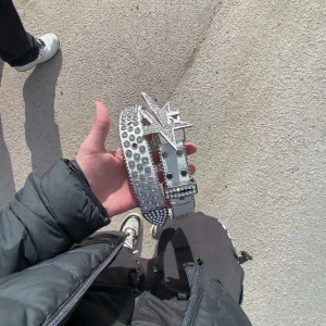 Vitt rhinestone bälte (lukamachain) - Säljer ett vitt bälte med stora silvriga nitar och glittrande detaljer. Bältet har ett unikt spänne i form av en stjärna med siffran 12, täckt av strass. Materialet är präglat för krokodilskinn-look och bältet är riktigt iögonfallande.