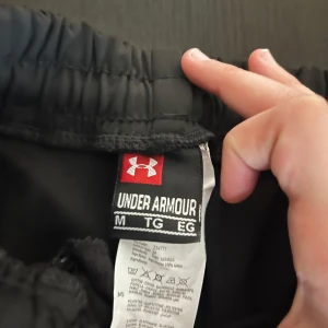 Svarta byxor o hoodie  från Under Armour - Svart hoodie o byxor  från Under Armour med elastisk midja och snörning. Klassisk sportig design med logga på vänster ben. Perfekta för träning eller avslappnade dagar. Tillverkade i ett lätt och funktionellt material.