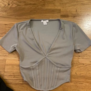 Ljusgrå korsettopp från NEW YORKER  - Trendig ljusgrå topp med korsettliknande detaljer och v-ringning. Toppen har korta ärmar och tight midja för en snygg siluett. Perfekt för att lyfta din outfit.
