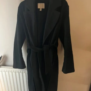 Svart kappa med bälte - Säljer en svart lång kappa med bälte i midjan som passar perfekt till höst och vår. Den har även slits på sidorna. Den är i storlek xs men passar perfekt som s