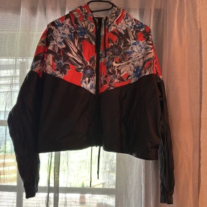 Blommig vindjacka från Nike - Säljer en färgstark vindjacka från Nike med blommigt mönster i blått, vitt och svart på röd botten. Jackan har huva, dragkedja och justerbar dragsko. Perfekt för dig som vill sticka ut med din stil!