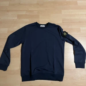 Mörkblå tröja från Stone Island - Säljer en stilren mörkblå tröja från Stone Island med deras ikoniska märke på ärmen. Tröjan har långa ärmar och en klassisk rund halsringning. Perfekt för en avslappnad och trendig look. Oanvän, lapparna vid magen är avklippta, pris kan diskuteras,
