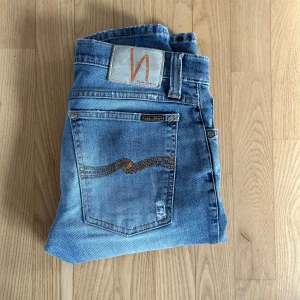 Ljusblå jeans från Nudie Jeans - Säljer ett par ljusblå jeans från Nudie Jeans med orange sömmar och signaturbroderi på bakfickorna. Normal passform och raka ben. Perfekta för en avslappnad stil. Är storlek 30/34