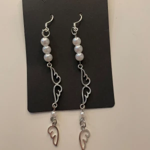 Långa örhängen med pärlor och vinge - Säljer ett par unika långa örhängen i silverfärgad metall med vita pärlor och dekorativa vinge-detaljer längst ner. Perfekta för dig som vill sticka ut med ett coolt och elegant smycke.