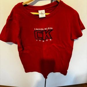 Röd t-shirt från Calvin Klein Jeans - Röd t-shirt från Calvin Klein Jeans med tryckt logga i vitt och svart på bröstet. Klassisk rund halsringning och korta ärmar. Perfekt för en avslappnad och trendig stil. Använd 1 gång