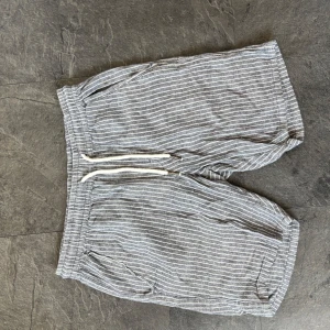 Gråvitrandiga linne shorts  - Snygga grå shorts med vita ränder och snörning i midjan. Shortsen har två sidofickor och två bakfickor. Perfekta för varma dagar och har en avslappnad passform.