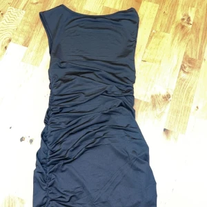Svart klänning - Snygg kort svart klänning med rynkade detaljer som ger en smickrande passform. Klänningen är ärmlös och lite öppen/djup i ryggen Materialet är mjukt och stretchigt för extra komfort. I storlek S 