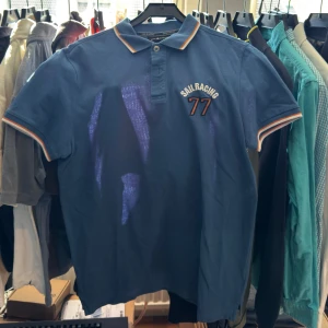 Blå piké från Sail Racing - Säljer en blå pikétröja från Sail Racing i storlek Large. Tröjan har korta ärmar, klassisk krage och knäppning framtill. Detaljer med orange och vit rand på krage och ärmslut samt broderad logga och siffran 77 på bröstet.