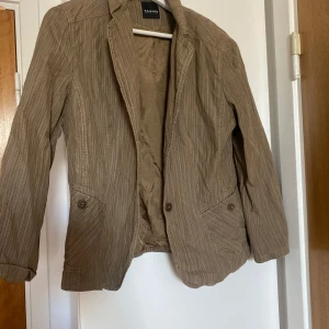 Beige kavaj från Taifun Collection - Säljer en beige kavaj från Taifun Collection med randigt mönster. Kavajen har två framfickor med knappar och klassisk krage. Materialet känns lätt och passar perfekt till vår och höst.