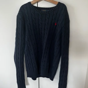 Mörkblå tröja från Polo Ralph Lauren - Säljer en mörkblå kabelstickad tröja från Polo Ralph Lauren med rund halsringning och röd broderad logga på bröstet. 