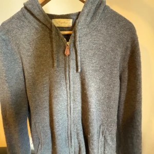 Grå kasmir zip hoodie ifrån Sayless - Säljer en stilren grå hoodie med dragkedja och huva. Perfekt för kyliga dagar. Den har en mjuk och bekväm passform med snörning vid huvan. Den är endast använd några gånger och ny pris 900kr