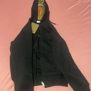 Svart hoodie från Burberry med dragkedja - Säljer en svart hoodie från Burberry med dragkedja och klassiskt rutigt foder i huvan. Tröjan har ribbade muddar och en stilren design med diskret logga på bröstet. Perfekt för dig som gillar exklusiva streetwear-plagg.