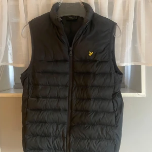 Svart dunväst från Lyle & Scott - Svart dunväst från Lyle & Scott med dragkedja framtill och klassisk gul logga på bröstet. Västen har en quiltad design och hög krage, perfekt för lager-på-lager-stil.