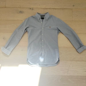 Ljusblå skjorta från Polo Ralph Lauren - Klassisk ljusblå skjorta från Polo Ralph Lauren i slim fit med button-down krage och broderad logga på bröstet. Skjortan har långa ärmar och vita knappar framtill. Perfekt för en stilren och tidlös look. Använd en gång. Skriv vid minsta fråga!