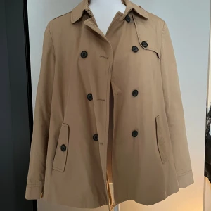 Beige trenchcoat med knappar Från Zara - Stilren beige trenchcoat med klassisk krage och svarta knappar framtill. Jackan är i storlek L men är mindre i storleken och passar mer s-m