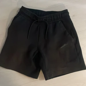 Svarta shorts från Nike med snörning - Svarta Nike-shorts med elastisk midja och snörning. Shortsen har sidofickor och en diskret Nike-logga framtill. Tillverkade i mjukt material som passar perfekt för träning eller vardag. Enkla och stilrena med sportig känsla.