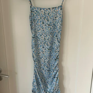 Blommig blå långklänning med smala axelband - Säljer en ljusblå långklänning med små vita och gula blommor. Klänningen har smala axelband och dragsko-detalj längs båda sidorna som ger en snygg rynkad effekt. Perfekt för sommarens varma dagar!