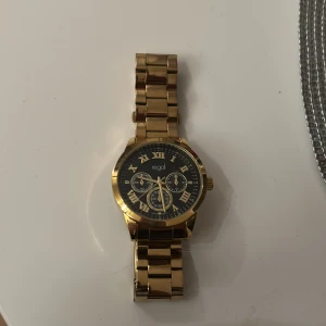 Guld klocka från regal - Stilren klocka från Regal med guldfärgad länk och svart urtavla. Klockan har romerska siffror, tre mindre urtavlor och klassisk design. Perfekt accessoar för dig som vill sticka ut med en elegant look.