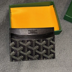 Svart korthållare  - Säljer en stilren svart korthållare med klassiskt mönster i vitt och grått. Korthållaren har flera fack för kort och är tillverkad i exklusivt skinn. Perfekt accessoar för dig som vill ha något både snyggt och praktiskt. Säljer pågrund av att jag inte använder den längre så det är dumt att ha den ligga och samla damm så då kan man sälja den vidare till en som faktiskt använder den. Pris kan diskuteras med snabb och smidig affär 