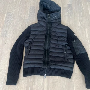 Svart hybridjacka med huva från Moncler - Säljer en svart Moncler-jacka med quiltad front och huva. Jackan har stickade ärmar och rygg, dragkedja framtill och fickor med knappar. Klassisk Moncler-logga på ärmen och praktisk ficka på ena ärmen. Perfekt för kyliga dagar.