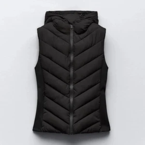 Svart dunväst med huva - Snygg svart dunväst med huva och dragkedja framtill. Västen har ett stilrent, vadderat mönster i sicksack och är figursydd för en modern look. Perfekt för lager-på-lager-stil under kyliga dagar. Den är även lite i storleken.