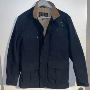 Mörkblå jacka från Barbour med brun krage - Klassisk mörkblå jacka från Barbour med kontrasterande brun krage i manchester. Jackan har flera fickor framtill, knappar och dragkedja. Perfekt för höst och vår. 