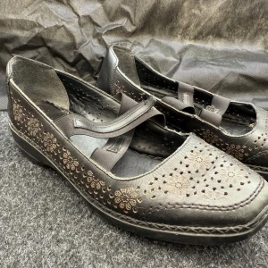 Svarta ballerinaskor med blommigt mönster - Säljer ett par svarta ballerinaskor i skinn med fina blommiga utskärningar och broderade detaljer. Skorna har elastiska band över foten för extra komfort och stil. Perfekta för dig som vill ha något både snyggt och bekvämt.