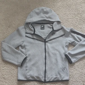 Nike tech fleece grå - Snygg grå tech fleece från Nike med svarta detaljer och dragkedja framtill. Jackan har huva, långa ärmar och två fickor på sidorna. Perfekt för en sportig och avslappnad stil.