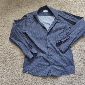 Blå mönstrad skjorta från Eton - Snygg blå skjorta från Eton med små vita och röda geometriska mönster. Skjortan har lång ärm, klassisk krage och knäppning framtill. Passformen är slim och materialet känns mjukt och svalt, perfekt till skjorta för dig som gillar stilren look.