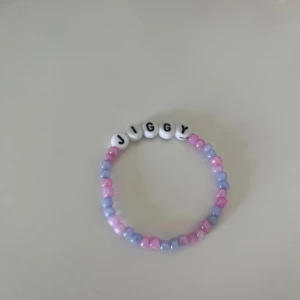 Pastellfärgat armband med bokstäver - Säljer ett sött armband med pastellfärgade pärlor i rosa och lila samt vita bokstavspärlor som stavar 'JIGGY'. Perfekt accessoar för att ge din stil en personlig touch! Hov1 inspirerat💘🫰🏻